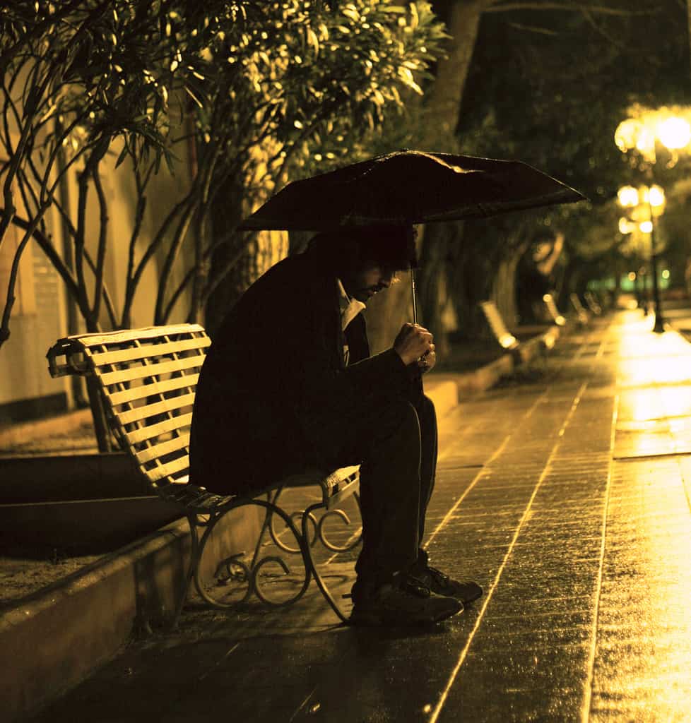 A person sitting alone on a park bench at night under an umbrella, reflecting on connection and solitude.