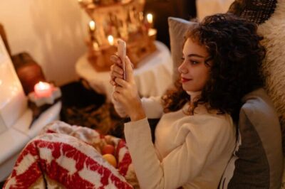 Woman relaxing at home during an online psychic video chat reading on her phone surrounded by soft candlelight and comfort.