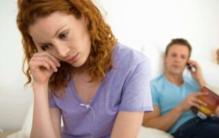 A woman sits thoughtfully on a bed, appearing emotionally distant as a man behind her looks preoccupied. The image represents emotional disconnection and the struggle to understand intimacy in relationships.