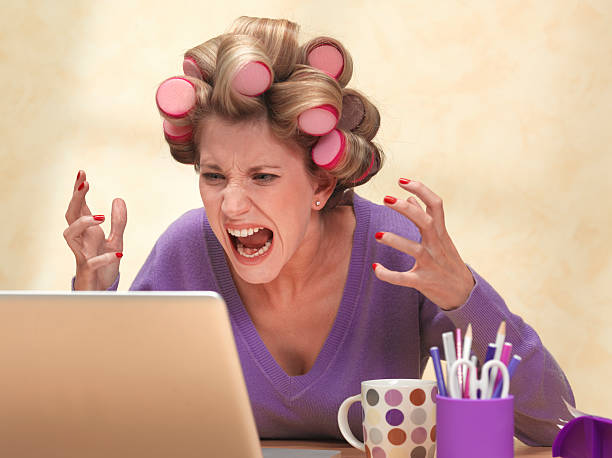 Frustrated woman shouting at her laptop with curlers in her hair, representing stress and the challenge of finding peace of mind in daily life.