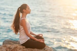 Woman sitting cross-legged by the ocean meditating peacefully at sunrise, symbolizing balance, mindfulness, and inner calm.