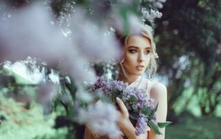 Woman standing quietly among lilac blossoms, holding flowers close with a serene expression, representing remembrance, gentle love, and spiritual connection.