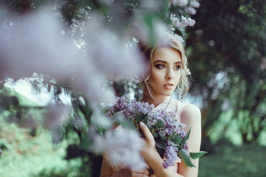 Woman standing quietly among lilac blossoms, holding flowers close with a serene expression, representing remembrance, gentle love, and spiritual connection.