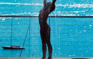 Woman standing under a sunlit outdoor shower beside a sparkling blue sea, arms lifted in joy — symbolizing renewal, cleansing, and self-love.