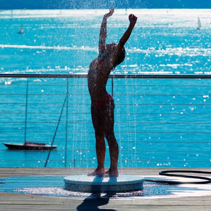 Woman standing under a sunlit outdoor shower beside a sparkling blue sea, arms lifted in joy — symbolizing renewal, cleansing, and self-love.