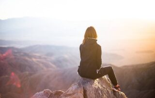 A woman sitting on a mountain rock at sunrise, reflecting quietly with courage and peace.